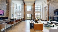 Located in the vibrant Taylor Street corridor of Houston, TX 77007, this elegantly designed community living space offers a striking blend of modern luxury and warm, inviting detail. The single-floor layout is defined by its soaring double-height ceilings that amplify the sense of openness throughout the room.

The living area serves as the centerpiece of the space, anchored by a floor-to-ceiling textured wood accent wall with a sleek, built-in linear fireplace and a wall-mounted television beneath a dramatic geometric wood-panel backdrop. A sculptural chandelier with cascading lights adds a refined focal point overhead.

Natural light pours in through expansive floor-to-ceiling black-framed windows and glass doors, offering views of the surrounding outdoor patio and landscaped courtyard. The light hardwood flooring, complemented by large-format tile near the entry, creates a seamless and sophisticated flow throughout.

Warm wood columns and custom built-in shelving units add architectural character, while a service counter with dark stone countertops is positioned for functionality. Multiple seating arrangements — featuring plush sofas, accent chairs in neutral and jewel tones, and stylish area rugs — provide ample space for gathering and relaxation. The overall ambiance is polished, contemporary, and welcoming.