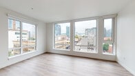 This single-floor unit is designed with a clean, contemporary aesthetic that emphasizes natural light and urban connectivity. The layout is anchored by a generously sized living area featuring large, floor-to-ceiling windows that wrap around the corner, framing sweeping views of the surrounding cityscape, including mid-rise and high-rise buildings, active streets below, and distant greenery. Light wood-tone flooring runs consistently throughout the unit, lending warmth and visual continuity across all spaces.

The open-plan kitchen is positioned adjacent to the living area, separated by a partial partition wall. It features flat-front white cabinetry extending to the ceiling, stainless steel appliances including a refrigerator, range, and microwave, as well as a textured tile backsplash and recessed lighting overhead. A tall, narrow window in the kitchen area introduces additional natural light.

A clean, white hallway connects the main living spaces to the private areas of the unit, where two bathrooms are found. Each bathroom includes a soaking tub with vertically tiled surrounds, a vanity with dark countertop, white cabinetry, and wood-tone flooring, maintaining a consistent modern finish throughout. The overall ambiance is bright, minimal, and urban.