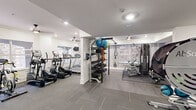 Located at 2900 Pharr Court South NW in Atlanta's prestigious Buckhead neighborhood, this well-equipped fitness center offers a comprehensive workout environment across a single floor. The space is finished with a clean, modern aesthetic, featuring crisp white walls accented by crown molding, recessed lighting, and ceiling fans that maintain a comfortable atmosphere throughout.

The cardio zone is dedicated to a row of treadmills and elliptical trainers positioned alongside windows that allow natural light and views of the surrounding exterior. A wall-mounted television provides entertainment during cardio sessions. An accent wall with a subtle wood-panel design adds a refined touch to this area.

The strength training section is equally well-appointed, housing a Cybex cable machine, a TRX suspension system, and a dedicated free weight area with a full rack of dumbbells, kettlebells, and medicine balls organized on a multi-tier storage rack. Additional equipment includes a rowing machine and a recumbent bike, catering to a variety of fitness routines.

Durable tile flooring with rubber mat overlays runs throughout, providing both safety and ease of maintenance. A water station is conveniently positioned near the entrance, and glass-paneled doors lead to adjoining common areas. The overall design is functional, clean, and thoughtfully organized to support a full range of fitness activities.