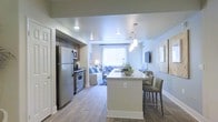 This single-floor space showcases a thoughtfully designed open-concept layout that integrates the kitchen, dining, and living areas into one cohesive environment. The kitchen is equipped with stainless steel appliances, including a full-sized refrigerator and a gas range, complemented by dark wood cabinetry that provides ample storage. A granite-topped kitchen island doubles as a breakfast bar with seating for multiple guests, making it a functional centerpiece for both meal preparation and casual dining. Pendant lighting hangs above the island, adding a modern decorative touch. The flooring throughout is a warm-toned hardwood, lending continuity and a sense of spaciousness across the open floor plan. The living area, visible from the kitchen, is bathed in natural light from a large window dressed with sheer curtains. Neutral-toned walls in soft blue-gray create a calm and inviting ambiance throughout. The space transitions effortlessly between functional and comfortable zones, making it well-suited for everyday living and entertaining alike.
