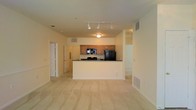 This single-floor space is designed with an open-concept layout that seamlessly connects the kitchen and living areas. The kitchen is outfitted with medium-toned wood cabinetry, a dark countertop peninsula, and black appliances including a refrigerator, built-in microwave, and range. The peninsula serves as a natural divider between the kitchen and the adjoining living space, making the area functional for both cooking and entertaining.

The living area features light beige carpeting that extends throughout, contributing to a warm and cohesive aesthetic. Overhead, a track lighting system provides directed illumination across the open space, complemented by a flush-mount ceiling fixture above the kitchen. The walls are finished in a neutral cream tone, enhancing the brightness of the interior.

Ventilation is provided through multiple ceiling and floor-level HVAC registers. A doorway visible on the left side of the space leads to additional rooms, suggesting a practical floor plan. The overall ambiance is clean, understated, and functional, suited for everyday living.