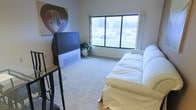 This single-floor space offers a straightforward and functional layout centered around a well-lit living area. The room features wall-to-wall carpeting that adds warmth underfoot, and a large picture window dressed with light curtains that allows generous natural light to fill the space while offering views of the surrounding outdoor area. The living area is furnished with a white slipcovered sofa, providing a clean and simple seating arrangement. A large rear-projection television is positioned across from the seating area, accompanied by a tall media stand and a potted indoor plant that adds a touch of greenery. A bold piece of wall art featuring a painted heart motif adds a personal and expressive element to the room. The neutral wall tones and understated décor create a calm, no-frills atmosphere throughout. Overall, the space presents a practical and livable environment with ample natural light and an open, uncluttered feel.