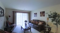 This single-floor residence offers a thoughtfully arranged living space designed for both comfort and functionality. The living room serves as the heart of the home, centered around a rich, dark leather sofa accented with decorative throw pillows in warm earth tones. A glass-topped coffee table with ornate wooden legs anchors the seating area, complementing the classic aesthetic of the space. Soft, neutral carpeting runs throughout, adding warmth underfoot. A large sliding glass door floods the room with natural light, framed by elegant burgundy and green drape panels with vertical blinds that allow for flexible light control. The door provides a seamless connection to the outdoors. Decorative wall art in complementary tones adds a personalized touch to the neutral walls, while a floor lamp in the corner contributes to the room's warm ambiance. The clean white ceiling with subtle architectural detailing maintains a bright, open feel throughout. Overall, the space presents a cozy and inviting atmosphere well-suited for everyday living and relaxed entertaining.