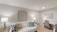 This renovated two-bedroom, one-bathroom single-floor residence offers a well-designed layout that maximizes comfort and functionality. The living room serves as a welcoming central space, featuring soft neutral walls, plush carpeting, and windows that invite natural light, creating a bright and airy atmosphere suited for both relaxing and entertaining.

The kitchen has been thoughtfully updated with white shaker-style cabinetry, light granite-look countertops, a deep double-basin stainless steel sink, and stainless steel appliances including a refrigerator and microwave. Wood-look flooring grounds the kitchen and extends into the adjacent dining area, which features a casual high-top table setup alongside a window and convenient entryway access.

Both bedrooms are carpeted and finished in soft gray tones, offering a calm, restful environment. Each room provides ample space for standard bedroom furnishings, with windows allowing natural light. The shared full bathroom is clean and functional, featuring a tub-shower combination, a vanity with a large mirror, and neutral finishes throughout.

Overall, the unit presents a cohesive, move-in-ready aesthetic defined by updated finishes, a neutral color palette, and a practical single-level floor plan.