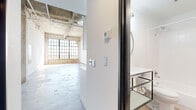 Located at 2400 S Ervay St in Dallas, Texas, this industrial-style loft showcases a thoughtfully converted open-plan layout with striking architectural character throughout. Soaring ceilings with exposed concrete soffits and visible ductwork and conduit lend an authentic warehouse aesthetic, while polished concrete floors anchor the space with understated texture and durability.

The expansive living area is bathed in natural light streaming through large, steel-framed grid windows set against a brick base wall — a hallmark of the building's industrial origins. A ceiling fan overhead adds functional comfort to the airy volume. A built-in steel ladder leads to an elevated loft area above, offering additional flexible space.

The combined dining and kitchen area features crisp white cabinetry, white quartz countertops, and a full suite of stainless steel appliances including a refrigerator, range, and dishwasher. Pendant industrial-style lights hang above, complementing the overall aesthetic seamlessly.

The bathroom is clean and modern, finished with light wood-look vinyl flooring, a floating metal-framed vanity, a round mirror, and a full tub-shower combo. A hallway with a closet provides practical storage. The unit also includes a dedicated entryway. Every detail of this space balances raw industrial character with modern, livable finishes.