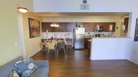 This single-floor space showcases an open-concept layout that connects the living, dining, and kitchen areas into one cohesive environment. The kitchen is well-appointed with rich dark-wood cabinetry offering generous storage, a stainless steel refrigerator, built-in microwave, and a light-toned countertop that wraps around to form a peninsula — ideal for casual dining or meal prep. A decorative tile backsplash adds visual texture to the cooking area.

The dining area sits at the heart of the space, anchored by a wooden table with upholstered chairs and illuminated by a classic chandelier with candle-style lighting, lending a warm, inviting atmosphere. An abstract wall painting nearby adds a touch of character to the dining nook.

Rich hardwood flooring runs throughout, unifying the entire floor plan. The living area, partially visible in the foreground, features a comfortable upholstered sofa. Neutral yellow-toned walls enhance the warmth of the space, while ceiling vents indicate a central climate control system. The overall ambiance is functional yet welcoming, with a balanced mix of modern appliances and traditional design elements.