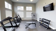 This fitness center offers a clean, functional environment designed to support a well-rounded workout routine. The space features soaring ceilings and a row of high-set windows that flood the room with natural light while providing views of the surrounding exterior, creating an open and energizing atmosphere. The walls are finished in a neutral gray tone, complemented by durable rubber flooring suited for high-traffic exercise use. A selection of cardio equipment lines one side of the room, including a treadmill and elliptical machines, making it easy to warm up or engage in sustained aerobic training. At the center of the space stands a cable machine, ideal for a wide variety of strength and functional training exercises. A flat utility bench provides a versatile surface for pressing movements and accessory work. Along the opposite wall, a tiered dumbbell rack holds a range of free weights, accommodating exercises at varying resistance levels. A wall-mounted flat-screen display adds an entertainment option for those looking to stay engaged during longer sessions. The overall layout is thoughtfully organized, offering clear circulation between equipment zones and a comfortable, uncluttered training environment.