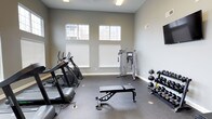 This fitness center offers a clean, functional environment designed to support a well-rounded workout routine. The space features soaring ceilings and a row of high-set windows that flood the room with natural light while providing views of the surrounding exterior, creating an open and energizing atmosphere. The walls are finished in a neutral gray tone, complemented by durable rubber flooring suited for high-traffic exercise use. A selection of cardio equipment lines one side of the room, including a treadmill and elliptical machines, making it easy to warm up or engage in sustained aerobic training. At the center of the space stands a cable machine, ideal for a wide variety of strength and functional training exercises. A flat utility bench provides a versatile surface for pressing movements and accessory work. Along the opposite wall, a tiered dumbbell rack holds a range of free weights, accommodating exercises at varying resistance levels. A wall-mounted flat-screen display adds an entertainment option for those looking to stay engaged during longer sessions. The overall layout is thoughtfully organized, offering clear circulation between equipment zones and a comfortable, uncluttered training environment.