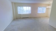 This single-floor space features a spacious, open living area defined by neutral cream-toned walls and wall-to-wall carpeting in a light grey hue, creating a clean and understated aesthetic. Two windows fitted with white horizontal blinds line the exterior wall, allowing generous natural light to fill the room while offering views of surrounding greenery. A baseboard heating unit is positioned beneath the larger of the two windows, suggesting an efficient and integrated heating solution. The textured ceiling complements the room's simple, functional design, while the open layout provides flexibility for a variety of furniture arrangements. The overall ambiance is quiet and airy, with the abundance of natural light and soft, neutral palette lending the space a calm and welcoming atmosphere. The room's proportions offer ample space for comfortable living, making it a versatile area well-suited to a range of uses.