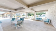 Located on Gusher Way in Lafayette, Indiana, this single-level outdoor amenity space is designed to serve as a premier community gathering destination. The covered pavilion is defined by its striking architectural elements, including a metal standing-seam roof supported by substantial natural stone columns and detailed coffered ceiling beams that add visual depth and structure overhead.

The interior of the pavilion is generously proportioned, offering multiple seating areas furnished with white-framed outdoor furniture accented by teal cushions. A standout feature is the full stone accent wall, which anchors a built-in fireplace topped by a mounted flat-screen television, creating an inviting focal point ideal for socializing in any season. French doors on either side of the fireplace wall connect the pavilion to adjacent interior spaces.

The open sides of the pavilion allow abundant natural light to filter in while maintaining unobstructed views of the surrounding landscaping and community grounds. A separate seating alcove along another stone wall provides a more intimate setting. Adjacent to the pavilion, a gated pool area with a dark metal perimeter fence offers a secure aquatic retreat, complete with lounge seating. A warm-toned timber pergola structure nearby further enhances the outdoor living experience, making this space a well-rounded and thoughtfully designed community amenity.