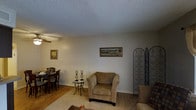 This single-floor space offers a welcoming and comfortable atmosphere with a thoughtfully arranged layout. The living area is anchored by a plush armchair and complemented by an ornate wrought-iron decorative screen, adding a touch of elegance to the room. Framed artwork adorns the neutral-toned walls, contributing to the warm and homey ambiance throughout. A small accent table with candleholders provides a charming focal point between the seating and dining areas. The dining area, visible to the side, features a classic dark wood table and chairs, creating an intimate setting for meals. The flooring transitions between hardwood in the dining area and carpet in the living space, offering both practicality and comfort. A textured ceiling adds subtle character to the room, while soft, diffused lighting maintains a calm and relaxed atmosphere. The overall design aesthetic leans toward a traditional and cozy style, with carefully chosen decorative elements that create a sense of warmth and livability throughout the space.