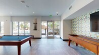 This single-floor amenity space is thoughtfully designed as a multifunctional recreation and social hub. The layout spans several interconnected zones, each catering to a distinct activity while maintaining a cohesive modern aesthetic throughout.

The game room area anchors one section of the space, featuring a full-size pool table and a shuffleboard table positioned on large-format stone-look tile flooring. Generous windows and glass doors along this zone flood the area with natural light and provide direct access to an outdoor terrace with views of a pool area beyond.

Adjacent to the game room, a dedicated arcade zone transitions to rich blue carpeting and includes vintage-style arcade cabinets, flat-screen gaming stations, a pinball machine, and bar-height tables with stools — creating an energetic entertainment hub.

A lounge seating area offers a more relaxed atmosphere, with a mustard-toned sofa, accent chairs, and a bold blue feature wall providing warmth and color contrast.

A dining nook features built-in bench seating backed by a striking geometric-patterned accent wall in teal, gold, and charcoal tones, complemented by round wood tables and light wood chairs.

A wet bar area showcases a marble countertop with built-in sinks, warm wood cabinetry, and a colorful blue-and-yellow tiled backsplash — a refined finishing touch to this well-appointed communal space.