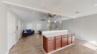 This single-floor space is designed around an open-concept layout that seamlessly connects the kitchen, dining, and living areas into one expansive, light-filled environment. The kitchen is a standout feature, equipped with rich dark cherry wood cabinetry, granite countertops, a classic subway tile backsplash, and ample storage throughout. A large central island with a granite surface provides generous counter space and doubles as a casual bar-height seating area, illuminated overhead by a row of stylish glass pendant lights.

The open living area extends naturally from the kitchen, showcasing vaulted ceilings that amplify the sense of space and airiness. Ceiling fans are positioned throughout to ensure comfort, and large windows dressed with white blinds invite abundant natural light while maintaining privacy. The hardwood-style flooring flows consistently across all areas, tying the space together with a warm, cohesive aesthetic.

A secondary lounge area, visible from the kitchen, offers additional flexibility and features its own vaulted ceiling, natural light from multiple windows, and space for both relaxed seating and recreational use. The neutral gray wall palette throughout creates a clean, modern backdrop that complements the warm wood tones of the cabinetry and flooring. Overall, this space offers a welcoming and functional environment well-suited for gatherings and everyday use.