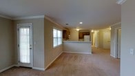 This single-floor residence offers a well-organized, open-concept layout designed for comfortable everyday living. The main living area is generously sized, featuring neutral wall-to-wall carpeting and warm beige walls that create a welcoming atmosphere. Crown molding runs along the ceiling, adding a classic architectural detail that elevates the overall aesthetic of the space.

The kitchen is positioned centrally and defined by a peninsula countertop that separates it from the living area, making it ideal for casual dining or meal preparation. Rich, warm-toned wood cabinetry lines the kitchen walls, complemented by a white refrigerator and built-in microwave. A window above the kitchen sink allows natural light to flow into the space.

A second window near the entry door further brightens the living area, contributing to an airy, open feel. Recessed lighting and a flush-mount ceiling fixture provide ample illumination throughout. A hallway visible in the background leads to additional rooms, offering a sense of depth and connectivity throughout the floor plan. The entry door and interior doors are framed with clean white trim, maintaining a cohesive and tidy finish. Overall, this space balances functionality with classic residential charm.