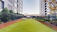 Located at 493 N Fifth St in Philadelphia's Northern Liberties neighborhood, this outdoor amenity courtyard offers a well-appointed urban retreat nestled between contemporary high-rise towers. The ground-level space is anchored by a generous stretch of vibrant artificial turf that provides an open, park-like atmosphere, ideal for recreation and relaxation.

Flanking the turf on both sides are robust Corten steel planter beds that add an industrial-modern aesthetic while housing a variety of lush shrubs, perennials, and manicured trees. Sleek black metal fencing runs the perimeter, providing a sense of enclosure and security without obstructing views of the surrounding architecture.

At one end of the courtyard, a covered outdoor bar area serves as the social focal point. Finished with a warm wood-paneled ceiling, a smooth concrete bar top, and bar-height seating, the structure offers a shaded gathering spot for residents. Wall-mounted lighting fixtures illuminate the space for evening use, enhancing its ambiance.

Concrete seating benches are thoughtfully positioned throughout the lawn area, offering casual places to sit and enjoy the open-air environment. The surrounding tower facades, characterized by large grid windows and a contemporary palette of light and dark materials, frame the courtyard and contribute to the cohesive modern design. This outdoor space strikes a balance between urban sophistication and approachable, functional comfort.