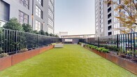 Located at 493 N Fifth St in Philadelphia's Northern Liberties neighborhood, this outdoor amenity courtyard offers a well-appointed urban retreat nestled between contemporary high-rise towers. The ground-level space is anchored by a generous stretch of vibrant artificial turf that provides an open, park-like atmosphere, ideal for recreation and relaxation.

Flanking the turf on both sides are robust Corten steel planter beds that add an industrial-modern aesthetic while housing a variety of lush shrubs, perennials, and manicured trees. Sleek black metal fencing runs the perimeter, providing a sense of enclosure and security without obstructing views of the surrounding architecture.

At one end of the courtyard, a covered outdoor bar area serves as the social focal point. Finished with a warm wood-paneled ceiling, a smooth concrete bar top, and bar-height seating, the structure offers a shaded gathering spot for residents. Wall-mounted lighting fixtures illuminate the space for evening use, enhancing its ambiance.

Concrete seating benches are thoughtfully positioned throughout the lawn area, offering casual places to sit and enjoy the open-air environment. The surrounding tower facades, characterized by large grid windows and a contemporary palette of light and dark materials, frame the courtyard and contribute to the cohesive modern design. This outdoor space strikes a balance between urban sophistication and approachable, functional comfort.