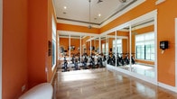 This fitness studio is designed to inspire activity through its bold aesthetic and well-appointed layout. The space is immediately distinguished by its striking orange walls, which are framed by crisp white crown molding and a contrasting gray trim band near the ceiling, lending the interior a polished yet energetic character. Light hardwood flooring runs throughout, reflecting natural light and contributing to the room's open, airy feel.

The cycling area is the centerpiece of the studio, housing a row of stationary spin bikes arranged for group classes. Full-length framed mirrors line the walls of this section, visually expanding the space and allowing participants to monitor their form during workouts. Ceiling fans provide ventilation overhead, and recessed lighting ensures consistent illumination throughout.

The open exercise area features tall windows along one wall, offering views of lush greenery outside and flooding the room with natural light. A sleek upholstered bench sits beneath the windows, providing a spot to rest or prepare. A wall-mounted flat-screen television and a built-in shelving nook add functional and informational elements to this area. Glass-paneled doors connect the spaces, maintaining an open flow while allowing for separation when needed.