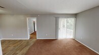 This single-floor unit is laid out across several well-defined spaces, each finished with neutral gray walls and clean white trim throughout. The living area is generously sized, featuring warm brown carpet and a sliding glass door fitted with vertical blinds that allow natural light to filter in while providing views of the surrounding greenery outside. A textured ceiling adds subtle dimension to the space.

Adjacent to the living area, a dining or entry space features wood-look plank flooring, providing a practical transition zone that connects directly to the kitchen. The galley-style kitchen is efficiently arranged with gray cabinetry, dark countertops, a stainless steel refrigerator, a range, and a window that brings in additional natural light. A fire extinguisher is mounted nearby for safety.

The bedrooms are carpeted and finished with the same neutral palette, each offering a ceiling fan or overhead light fixture and windows with blinds. One bedroom includes access to a walk-in closet. The bathroom is equipped with a gray vanity, granite-style countertop, a large mirror, a well-lit vanity fixture, and wood-look flooring, offering a clean and functional layout throughout.
