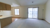 This single-floor space offers a clean, open layout designed for comfortable everyday living. The main living area is finished with wall-to-wall carpeting and bathed in natural light from two generously sized windows, including a large sliding glass door that opens directly onto an outdoor deck, providing a seamless connection between interior and exterior spaces. The sliding door is dressed with sheer curtains that soften the incoming light while maintaining a bright and airy atmosphere. The warm, neutral wall tones throughout the living area create a welcoming and versatile backdrop.

Adjacent to the living area, the space transitions into a kitchen zone distinguished by smooth hardwood-style flooring, offering a practical and visually appealing contrast to the carpeted areas. A countertop overhang hints at a breakfast bar or pass-through design, enhancing the open feel between the kitchen and living spaces. Overhead ventilation is present, and recessed ceiling vents ensure consistent climate control throughout. The overall design is functional and understated, with wood-trimmed windows adding a classic, grounded aesthetic. The layout is ideal for those seeking a straightforward, light-filled living environment.