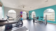 This fitness center in Robinsonville, Mississippi, offers a compact yet well-appointed exercise space designed to support a well-rounded workout routine. The single-room layout is finished with durable dark flooring, a textured ceiling, and a ceiling fan with integrated lighting that helps maintain a comfortable environment during workouts. A bold teal accent wall serves as the room's visual centerpiece, adorned with the motivational phrase "Train Hard You Can," adding energy and character to the space. Multiple arched windows line the walls, flooding the room with natural light and providing views of the surrounding outdoors. The facility is equipped with a variety of cardio machines, including treadmills, an elliptical, and a stationary bike, alongside cable and weight machines for strength training. Exercise balls in multiple sizes are also available for functional fitness routines. A wall-mounted television provides entertainment during workouts. French doors offer direct access to the exterior, enhancing ventilation and connectivity to outdoor areas. Recessed lighting supplements the natural light for evening use. The overall ambiance is bright, clean, and motivating, making this fitness center a practical and inviting amenity for residents of the Robinsonville community.