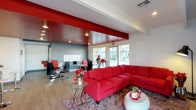 This single-floor space makes an immediate visual impact with its bold, cohesive red-and-white design aesthetic. The main area is thoughtfully divided into two distinct zones that flow seamlessly into one another. The lounge area anchors the space with a large red sectional sofa, complemented by a round glass-top side table and a contemporary white ottoman, all set atop a patterned area rug over warm hardwood-style flooring. Fresh floral arrangements in red tones add vibrant decorative accents throughout.

A defining architectural feature is the deep red ceiling panel that extends over the workstation zone, creating a striking visual contrast against the white perimeter ceiling. This area is furnished with sleek black desks and red ergonomic chairs, suggesting a professional or client-facing workspace. A large whiteboard mounted on the gray accent wall serves as a practical focal point in this section.

Large windows along the rear wall flood the interior with natural light, offering views of the outdoors and enhancing the open, airy feel of the space. Recessed lighting supplements the natural light, ensuring the environment remains bright and welcoming. The overall ambiance is energetic and modern, with the consistent use of red as a unifying design element throughout.