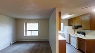 This single-floor space offers a straightforward, functional layout with clearly defined living and kitchen areas. The living room is finished with neutral wall-to-wall carpeting and plain white walls, creating a blank canvas well-suited for personalization. A textured popcorn ceiling runs throughout, adding a characteristic finish consistent with the unit's construction era. Baseboard heaters line the perimeter walls, providing an efficient heating solution. A window along the living room wall allows natural light to filter into the space, contributing to a bright, open feel.

The kitchen is partially separated from the living area by a half-wall partition, maintaining a sense of openness while defining each zone. It features medium-toned wood cabinetry, a white refrigerator, a white electric range, and a light-colored countertop. A second window in the kitchen area enhances natural light and ventilation. Overhead fluorescent lighting provides ample task illumination. An adjacent area transitions to hardwood-style flooring, offering a visual contrast to the carpeted living spaces. Overall, the unit presents a clean, utilitarian aesthetic suited to practical, everyday living.