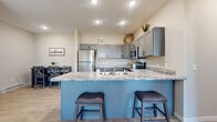 This single-floor home combines modern design with functional living across a well-organized layout. The kitchen serves as the centerpiece of the space, showcasing a bold blue peninsula island topped with striking veined granite countertops and seating for two. Gray shaker-style cabinetry lines the walls, complemented by stainless steel appliances including a refrigerator, microwave, and range. Recessed lighting illuminates the kitchen and flows into the adjacent dining area, which connects seamlessly to the rest of the home. The living room features light wood-look flooring, a ceiling fan, and a front entry door flanked by a large window that fills the room with natural light. Two bedrooms are finished with plush carpeting and generously sized windows offering neighborhood views. The primary bedroom includes a ceiling fan, while the secondary bedroom features a sliding closet with built-in shelving. Both rooms offer a calm, comfortable atmosphere. The attached two-car garage is drywalled with a concrete floor and an automatic opener, providing ample storage and parking capacity. Throughout, neutral wall tones and clean finishes create a cohesive, welcoming environment.