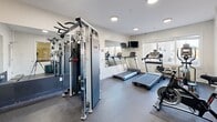 Located at 255 East Grand Avenue in Rahway, New Jersey, this fitness center is a well-appointed facility designed to support a comprehensive workout routine. The space features durable rubber flooring throughout and is bathed in natural light from multiple large windows that offer views of the surrounding exterior. Recessed and flush-mount ceiling lighting supplements the natural light, ensuring a bright and energizing environment at any time of day.

The cardio area is equipped with multiple treadmills and an elliptical machine, positioned to take advantage of the natural light from the windows. A wall-mounted flat-screen television is also present for entertainment during workouts. A multi-functional cable weight station anchors the strength training section, accompanied by a full rack of hex dumbbells and a yoga mat for versatile training options. Large wall mirrors expand the sense of space and allow for proper form checks during exercise.

The walls are finished in clean white, complemented by a bold motivational "DON'T QUIT" wall graphic that adds character and energy to the room. Posted fitness center rules and hours ensure an organized, shared-use environment. The space is accessed through a paneled white door and connects to adjacent building areas, making it a convenient and fully integrated amenity for residents at this Rahway address.
