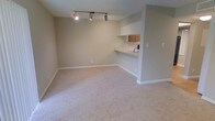 This single-floor space is designed with an open and functional layout that maximizes its available square footage. The main living area is finished with neutral-toned wall-to-wall carpeting that lends a clean, understated aesthetic throughout. A track lighting fixture mounted on the ceiling provides adjustable, directional illumination, offering both practicality and a subtle modern touch.

A defining architectural feature is the built-in pass-through opening that connects the living area to the kitchen. This cutout is accented by a countertop ledge, which can serve as a breakfast bar or convenient pass-through surface. Above the opening, a wall-mounted shelf adds additional storage or display space. The kitchen area, visible through the pass-through, includes warm-toned wood cabinetry and a sink, maintaining a cohesive and efficient workspace.

The walls are finished in a consistent warm beige tone, complementing the light carpeting and contributing to an overall neutral, move-in-ready palette. White baseboards frame the perimeter of the room cleanly. The space presents a straightforward, well-maintained interior suited for everyday living.