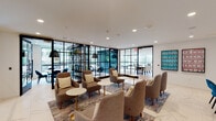 This thoughtfully designed single-floor amenity space blends contemporary aesthetics with versatile functionality. The layout is anchored by a spacious lounge area featuring polished marble-effect tile flooring, plush upholstered armchairs in warm tones, and low marble-top accent tables arranged atop a vibrant area rug. Curated artwork adorns the walls, adding pops of color against the clean white backdrop. A dining nook with banquette seating, teal accent chairs, and white tabletops provides a casual gathering spot, while a glass railing and staircase add an open, airy architectural element to the transitional corridor. Black-framed steel and glass partitions divide the space into distinct zones while maintaining visual connectivity and allowing natural light to flow throughout. Private rooms enclosed by these partitions offer more intimate settings for meetings or focused work. A dedicated game room with herringbone hardwood flooring features a ping-pong table and additional dining-style seating, with large windows offering views of the surrounding streetscape. The kitchenette is appointed with sleek flat-front white cabinetry, a herringbone tile backsplash, matte black hardware and open industrial shelving above the countertop. Recessed lighting throughout ensures a bright, welcoming atmosphere across every area of the space.