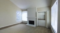 This single-floor space is defined by a clean, understated aesthetic that balances functional design with comfortable living. The main living area draws immediate attention with its vaulted ceiling, which adds a sense of openness and volume to the room. A prominent white brick fireplace serves as the focal point, extending nearly floor to ceiling with a slatted surround detail that adds subtle architectural texture. Neutral-toned carpet covers the main floor area, while a tile border runs along the perimeter, adding a low-maintenance and visually grounding element to the space.

Natural light fills the room through a corner window arrangement fitted with horizontal blinds, offering views of mature outdoor greenery and providing a pleasant connection to the exterior. The light walls and white trim throughout reinforce a bright, airy atmosphere.

Adjacent to the living area, a recessed nook houses what appears to be a wet bar or compact kitchenette, complete with white cabinetry, a small countertop, and a sink — a convenient and practical feature. A louvered door nearby suggests additional storage or utility access. Overall, the space presents a well-organized, move-in-ready layout with timeless, neutral finishes throughout.