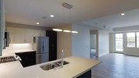 This single-floor unit showcases a contemporary open-concept design that seamlessly integrates the living and kitchen areas. The kitchen is a standout feature, equipped with stainless steel appliances including a French door refrigerator, a range, and a microwave. Dark cabinetry on the lower level contrasts with lighter upper cabinets, creating a visually balanced aesthetic. A spacious kitchen island with a double stainless steel sink and a sleek pendant light fixture serves as a functional centerpiece, ideal for both meal preparation and casual dining.

The flooring throughout is consistent wood-style planking, adding warmth and continuity to the space. Recessed lighting in the kitchen and ceiling-mounted fixtures provide ample illumination. The open living area offers a generous layout with neutral white walls, allowing flexibility for various furnishing arrangements.

A dedicated in-unit laundry area is conveniently located near the main living space, housing a stacked washer and dryer. A hallway visible from the main area leads to additional rooms, suggesting a well-organized floor plan. HVAC vents are integrated into the ceiling, ensuring efficient climate control throughout the unit.