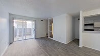 This single-floor residence offers a clean, modern aesthetic throughout, highlighted by consistent wood-look flooring that flows seamlessly from room to room. The open living area is a welcoming focal point, featuring a textured ceiling, crisp white walls, and a sliding glass door that leads to a private balcony, allowing natural light to fill the space. A built-in shelving unit serves as a subtle divider, adding functional storage while maintaining the open feel of the layout. The kitchen is efficiently designed with white cabinetry, granite-style countertops, a classic white subway tile backsplash, and black appliances including a range, refrigerator, and dishwasher. A double-basin stainless steel sink with a modern faucet completes the cooking space. The bedroom is generously proportioned with clean white walls and direct access to two doors, providing privacy and convenience. The bathroom is a standout feature, offering a wide vanity with a granite-style countertop, an undermount sink, white cabinetry, and a large window dressed with blinds that invites natural light. An adjoining walk-in closet with a built-in rod and shelf provides ample storage. The overall ambiance is bright, neutral, and move-in ready.
