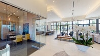 This single-floor clubhouse is designed with a contemporary aesthetic, combining open-plan living spaces with thoughtfully defined functional zones. The grand entrance lobby immediately impresses with soaring ceilings, large arched windows, and floor-to-ceiling glass doors that flood the space with natural light. Elegant pendant chandeliers and recessed lighting complement the bright, airy atmosphere, while large-format tile flooring and clean architectural lines reinforce the modern design throughout.

The main lounge area features plush seating, natural wood coffee tables, and vibrant area rugs, creating an inviting social environment. Floor-to-ceiling black-framed windows frame lush outdoor palm tree views, seamlessly connecting the interior to the surrounding landscape. Adjacent to the lounge, a sophisticated kitchen and bar area showcases warm wood cabinetry, quartz countertops with a herringbone tile backsplash, open wooden shelving, and stainless steel appliances—ideal for social gatherings.

A glass-enclosed workspace provides a private yet visually connected environment for productivity. The fitness center is generously sized, featuring hardwood-style flooring, large windows overlooking the outdoors, cardio equipment, free weights, and a motivational graphic accent wall. A glass-enclosed children's play area is thoughtfully integrated within the fitness space, ensuring supervision remains easy.