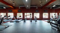 This fitness center is a thoughtfully designed, single-floor workout facility that offers a comprehensive range of exercise equipment and a vibrant, energizing atmosphere. The space is defined by its bold color palette, featuring deep red accent walls and orange geometric color blocks that create a dynamic and motivating environment.

The cardio zone is lined along a row of large windows, which flood the area with natural light and provide views of the surrounding outdoor greenery. This section houses multiple treadmills, elliptical trainers, and stationary bikes, all equipped with individual display screens. Ceiling fans and exposed industrial-style ductwork add to the open, high-energy feel of the space.

On the opposite side, the strength training area features a textured decorative wall panel in warm tones, serving as an eye-catching focal point. This zone is outfitted with cable machines, a weight bench, a dumbbell rack, stability balls, and a digital fitness kiosk for guided workouts.

Full-length mirrors span an entire wall, visually expanding the space and allowing users to monitor their form throughout their workouts. The durable rubber flooring provides both safety and functionality across the entire facility. Recessed lighting complements the natural light, ensuring the space is well-lit at all times. Overall, the fitness center delivers a professional and well-appointed workout experience.