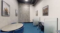 Located at 307 SW 5th St in Fort Lauderdale, FL, this coworking office space offers a thoughtfully designed environment ideal for productivity and collaboration. The single-room space makes efficient use of its footprint, accommodating both collaborative and individual work needs.

The interior is defined by a striking industrial aesthetic, with exposed black ceiling beams and ductwork contrasting against crisp white walls. A bold geometric-patterned feature wall anchors one end of the room, adding visual depth and character. The vivid blue carpet throughout grounds the space with a pop of color that energizes the environment.

The layout is divided into two functional zones: a casual meeting area centered around a round white table with modern blue bucket chairs, and a dedicated workstation section furnished with ergonomic task chairs and frosted glass privacy dividers. Curated framed artwork adorns the walls, lending a creative, gallery-like atmosphere.

Entry is through full-height black-framed glass doors with frosted branding decals, providing both transparency and a polished professional appearance. Recessed ceiling lighting ensures consistent, even illumination throughout the space. The overall design balances open visibility with focused work areas, making it a versatile and well-appointed office environment suited for small teams or individual professionals.