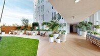 This expansive amenity space is thoughtfully designed to blend indoor comfort with outdoor living, offering a generous footprint that accommodates a diverse range of activities and social settings. The modern aesthetic is defined by clean white facades, light wood herringbone flooring, and an abundance of natural light throughout.

The outdoor courtyard serves as a central gathering area, featuring manicured turf lawns, lush tropical and native plantings in sculptural white planters, and casual seating including wooden lounge chairs and woven accent chairs. The open-air design is framed by the building's multi-story contemporary architecture, with glass balcony railings visible on upper levels.

An adjacent covered terrace transitions seamlessly between indoors and out, furnished with striped upholstered benches, concrete accent tables, and tall tropical plants. Floor-to-ceiling glass barriers open the space to sweeping skyline views, while vertical slatted wall panels add architectural texture.

The state-of-the-art fitness center is fully outfitted with treadmills, stationary bikes, kettlebells, punching bags, resistance bands, a trampoline, and large-screen displays. Industrial-style exposed ceilings with linear lighting give the gym a modern, energetic atmosphere. Folding glass doors connect the fitness center directly to the outdoor turf area, allowing for seamless indoor-outdoor workouts.