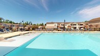This single-level community amenity space features two distinct areas designed for recreation and wellness. The outdoor pool area is the centerpiece of the community's leisure offerings, showcasing a generously sized, crystal-clear swimming pool with a shallow entry section and a surrounding deck lined with lounge chairs and classic pole-mounted light fixtures. The pool area is enclosed by black iron fencing, with mature trees and residential buildings visible in the background beneath open blue skies.

The indoor fitness center is thoughtfully designed with a teal and sage green color-blocked accent wall bearing a bold "fit·ness" graphic. The space is flooded with natural light through large floor-to-ceiling windows and features light hardwood-style flooring throughout. Equipment includes Matrix-brand treadmills, ellipticals, stair climbers, stationary bikes, and a spin cycle, as well as free weights, resistance bands, exercise balls, and yoga mats. A wall-mounted TV and ceiling fan add to the comfort of the space. A unique garage-style door opens directly to an outdoor pergola terrace, seamlessly blending indoor and outdoor workout environments. A small refreshment station and beverage cooler are also present near the entrance.