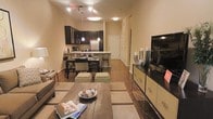 This single-floor residence showcases an open-concept layout that integrates the living, dining, and kitchen areas into one cohesive, flowing space. The living area is anchored by a rustic wooden coffee table paired with neutral-toned seating and complemented by a pair of olive green ottomans, adding a touch of color. A flat-screen television is mounted on a sleek, decorative media console along the right wall, which features stylish accents and display pieces.

The dining area transitions naturally from the living space, featuring a dark-toned dining table with matching chairs and patterned cushions, set atop warm hardwood flooring that runs throughout the entire space. Pendant lighting above the kitchen counter adds ambient illumination.

The kitchen is appointed with dark espresso cabinetry, a stainless steel refrigerator, and a raised breakfast bar with seating. Track lighting on the ceiling provides practical overhead illumination, enhancing the warm, inviting ambiance. A hallway visible in the background leads to additional areas of the residence. The overall atmosphere is modern, cozy, and well-appointed.
