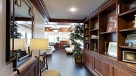 This single-story space showcases refined traditional design through its prominent use of rich, dark wood millwork that frames the ceilings, walls, and built-in shelving throughout. The main living area draws the eye down a well-proportioned corridor flanked by floor-to-ceiling built-in bookcases with display shelving, ideal for books, art objects, and decorative accents. Dark hardwood flooring runs continuously through the space, anchoring the warm, classic aesthetic. A coffered ceiling detail adds architectural depth and visual interest overhead, while recessed and pendant lighting provide layered illumination. At the far end of the space, a fireplace serves as a natural focal point, surrounded by a traditional mantel and framed by seating. A tall indoor tree introduces a natural, organic element that softens the richness of the woodwork. Natural light filters in through windows visible in the background, brightening the interior. The overall ambiance is one of traditional elegance and comfort, with careful attention to craftsmanship and detail throughout every element of the design.