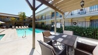 This outdoor communal area is centered around a generously sized swimming pool with clear turquoise water, complemented by an adjacent spa. The pool deck is lined with white lounge chairs and a shaded umbrella station, providing ample space for relaxation under open skies. The courtyard is framed on multiple sides by a two-story building featuring iron-railed balconies, stucco exteriors, and arched architectural accents, creating an enclosed and private atmosphere.

A standout feature of the space is the wood-framed pergola structure, which provides partial shade over a dedicated outdoor dining and lounge area. Beneath the pergola, a wicker dining table with cushioned chairs offers a comfortable setting for gatherings, while a built-in BBQ grill station nearby adds functional outdoor entertaining capability. A hanging lantern-style light fixture adds a decorative touch to the covered area.

The pergola's slatted roof allows filtered natural light to pass through, and climbing vines add a natural, lush element to the structure. Manicured hedgerows and landscaped greenery border the pool area, enhancing privacy. The overall ambiance is bright, sun-drenched, and well-suited for outdoor leisure and social activity.