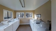 This shared laundry facility is designed with practicality and convenience in mind. The room is spacious enough to accommodate two rows of full-sized, top-loading washing machines and dryers arranged along opposing walls, providing ample capacity for multiple users. The warm beige-painted walls create a neutral, welcoming atmosphere, while the smooth white ceiling with overhead fluorescent lighting ensures the space is well-lit throughout. Two large windows along the back wall invite natural light into the room and offer views of the surrounding building exterior, adding an airy quality to the functional space. The flooring is finished with a durable, low-pile carpet in a neutral tone, contributing to a quiet and comfortable environment. An informational board is mounted near the machines, offering guidance and notices for users. The room's clean, organized layout makes navigating and using the facilities straightforward. Overall, this laundry facility is thoughtfully arranged to serve the practical needs of its users efficiently, with a tidy and maintained appearance that reflects attentive upkeep.