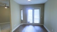 This single-floor space offers a clean, functional layout with a warm and inviting ambiance. The interior is finished with rich, dark hardwood flooring that runs throughout, providing a classic and durable foundation that complements the soft, neutral sage-green wall tones. A pair of French doors serves as a central architectural feature, flooding the space with generous natural light and offering a direct connection to the outdoors. Horizontal blinds on both the French doors and the accompanying side window allow for flexible light and privacy control. The ceiling is fitted with a ceiling fan and light fixture, ensuring year-round comfort and practical illumination. Clean white baseboards and door trim provide a crisp contrast against the muted walls, contributing to a well-finished aesthetic. The open, uncluttered layout makes the space highly adaptable for a variety of uses. Overall, the space strikes a balance between functional design and comfortable livability, with natural light and quality finishes as its defining characteristics.