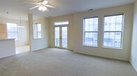 This two-floor space features a generously sized main living area finished with wall-to-wall neutral carpeting that lends a clean, cohesive feel underfoot. The room is bathed in natural light, courtesy of multiple double-hung windows positioned along the walls, as well as a set of elegant French doors topped with a transom window. The French doors open to a small exterior balcony, offering views of the surrounding urban streetscape. Soft, cream-toned walls complement the light streaming in from multiple directions, creating a warm and inviting ambiance throughout. A ceiling fan with an integrated light fixture is centrally mounted, providing both air circulation and ambient lighting. A pass-through cutout in one of the walls hints at an adjacent kitchen or secondary room, adding functional connectivity within the layout. The overall design is straightforward and practical, with clean lines and neutral finishes that provide a versatile canvas for a variety of interior styles. The multi-floor layout offers additional living space beyond what is visible in the main area.