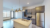 This thoughtfully designed one-bedroom, one-bathroom unit offers an efficient and functional layout across a single floor. The kitchen serves as a standout centerpiece, featuring a large island with a granite countertop and an integrated sink, taupe shaker-style cabinetry, a mosaic tile backsplash, and a full suite of stainless steel appliances including a refrigerator, electric range with microwave, and dishwasher. Striking brushed-metal industrial pendant lights hang above the island, adding a modern, urban aesthetic. The kitchen flows seamlessly into the living area, which is finished with warm wood-look plank flooring that enhances the open feel of the space. The bedroom is carpeted for comfort and features a ceiling fan, neutral toned walls, and a large window with a roller shade that frames views of the surrounding neighborhood and fills the room with natural light. The bathroom is finished with wood-look tile flooring, a dark-toned vanity with a marble-style countertop, a framed mirror, and modern light fixtures. A dedicated closet provides practical storage. Bold dark-painted interior doors and trim add a contemporary contrast throughout the unit, contributing to its cohesive modern design.
