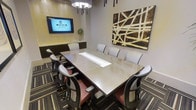 This single-floor conference room is designed to facilitate professional meetings and collaborative gatherings in a polished, contemporary setting. At the heart of the space sits a large, rectangular meeting table finished in a light wood tone, surrounded by ergonomic chairs upholstered in neutral tones with subtle pink accents — comfortably accommodating a sizeable group. A wall-mounted flat-screen display positioned at the head of the room ensures seamless presentations and video conferencing capabilities. The room's design aesthetic is refined and modern, with textured wall panels in muted tones providing an elegant backdrop. A striking large-format abstract artwork in gold, white, and dark brown tones adorns one wall, adding a creative focal point to the space. Recessed and pendant lighting fixtures cast a warm, even glow across the room, enhancing the professional atmosphere. The patterned carpeting in muted stripes of brown and gold grounds the space with a sophisticated touch. A built-in counter or credenza along one wall offers additional functionality for refreshments or materials. Overall, the space strikes a balance between business-ready utility and thoughtful interior design.