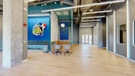 Located at 222 Canal St in New Haven, Connecticut, this single-floor commercial lobby offers a generously proportioned, open-plan space designed with a bold and contemporary aesthetic. Light hardwood flooring spans the majority of the space, complemented by colorful area rugs that define distinct zones within the layout.

The design makes a strong visual statement through vibrant cobalt and teal accent walls adorned with large-scale original artwork, creating a dynamic and energetic atmosphere. A curved, wood-paneled reception desk anchors the central area, paired with modern task chairs and a blue pegboard backdrop. Nearby, a striped sectional sofa provides a welcoming lounge area, while a striking spiral red sofa serves as a sculptural focal point in the open gathering zone.

Soaring ceilings with exposed ductwork and structural beams lend the space an industrial character, softened by warm pendant globe lighting. Floor-to-ceiling glass windows along one elevation flood the interior with natural light and offer open views of the surrounding New Haven streetscape. Glass-walled private office or conference enclosures line one side of the space, maintaining transparency while providing functional separation. A dedicated elevator lobby, finished with a teal ceiling and pendant lights, ensures smooth vertical access throughout the building.