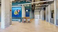 Located at 222 Canal St in New Haven, Connecticut, this single-floor commercial lobby offers a generously proportioned, open-plan space designed with a bold and contemporary aesthetic. Light hardwood flooring spans the majority of the space, complemented by colorful area rugs that define distinct zones within the layout.

The design makes a strong visual statement through vibrant cobalt and teal accent walls adorned with large-scale original artwork, creating a dynamic and energetic atmosphere. A curved, wood-paneled reception desk anchors the central area, paired with modern task chairs and a blue pegboard backdrop. Nearby, a striped sectional sofa provides a welcoming lounge area, while a striking spiral red sofa serves as a sculptural focal point in the open gathering zone.

Soaring ceilings with exposed ductwork and structural beams lend the space an industrial character, softened by warm pendant globe lighting. Floor-to-ceiling glass windows along one elevation flood the interior with natural light and offer open views of the surrounding New Haven streetscape. Glass-walled private office or conference enclosures line one side of the space, maintaining transparency while providing functional separation. A dedicated elevator lobby, finished with a teal ceiling and pendant lights, ensures smooth vertical access throughout the building.