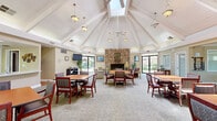 Located at 35 W Clover St in Woodland, California, this single-story community clubhouse offers a generously sized, well-appointed gathering space designed for comfort and shared living. The architectural centerpiece is the expansive main living area, crowned by soaring vaulted ceilings with exposed white-painted beams and multiple skylights that flood the interior with natural light. A dramatic floor-to-ceiling stone fireplace anchors the lounge area, which is furnished with comfortable seating arrangements ideal for relaxed conversation. Large windows and glass doors line the perimeter, providing abundant views of the surrounding outdoor greenery. The main hall accommodates multiple dining and activity tables, offering ample space for community gatherings. A dedicated game room extends from the main area and features a full-size pool table, additional seating, a bookshelf library nook, and access to a refreshment station. The hallways connect seamlessly to a well-equipped laundry room outfitted with multiple full-size washers and dryers, utility sinks, and folding counters beneath warm-toned walls. A tiled full bathroom with accessible fixtures, including grab bars and a urinal, serves the facility. Throughout, neutral carpeting, indoor greenery, and warm lighting create an inviting and functional communal environment.