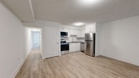 Located at 1171 Lansdowne Ave in Toronto's Dovercourt-Wallace Emerson neighbourhood, this 2-bedroom, 2-bathroom lower unit offers a clean, modern interior with a practical layout across a single floor. Light wood laminate flooring runs throughout, complementing the crisp white walls and creating a bright, cohesive aesthetic.

The open-concept kitchen and living area forms the heart of the unit. The kitchen is equipped with stainless steel appliances, including a full-size refrigerator and electric range, and features white cabinetry. The adjoining living space is generously proportioned, with windows that allow natural light to filter in.

Both bedrooms are well-sized, with windows providing garden-level views of surrounding greenery. The primary bedroom includes a double-door closet for ample storage. Each room maintains the consistent light flooring and neutral palette found throughout the unit.

The two bathrooms are finished with classic white subway tile. One features a full soaker tub with an overhead shower, while the second offers a standalone walk-in shower with grey hexagonal floor tile and chrome fixtures — both clean and functional in design.

Two hallways connect the rooms efficiently, and the unit includes an accessible electrical panel. The overall ambiance is fresh and move-in ready, with a modern sensibility suited to comfortable urban living.